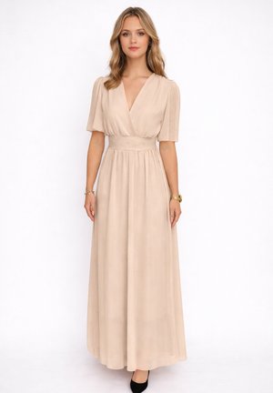 Femme portant une robe maxi beige à manches courtes et col en V, taille cintrée et chaussures noires, debout devant un fond blanc uni.