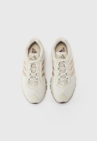 Lys beige idrettssko med mesh- og syntetisk overdel, med tre brune striper, snøring og en teksturert såle.