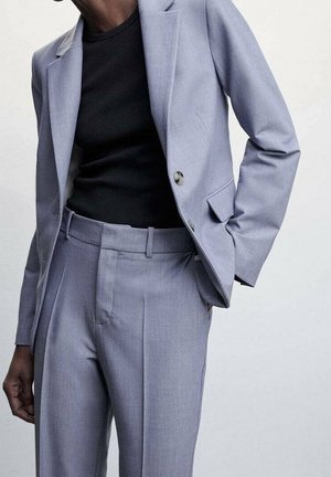 Personne portant un costume gris clair avec un blazer à un bouton et un pantalon assorti par-dessus un haut noir ajusté, posant avec une main dans la poche.