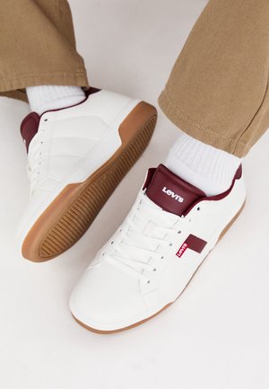 Hvide sneakers med brune såler og bourgogne accenter båret med hvide sokker og beige bukser, der viser brandlogoer på tunge og side.