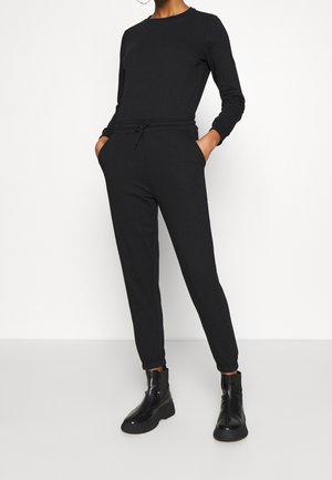 Person trägt einen figurbetonten schwarzen Overall mit langen Ärmeln und Kordelzug in der Taille sowie schwarze Stiefeletten, die Hände in den Taschen, steht auf einem weißen Boden.