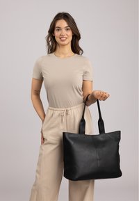 Zwarte leren tote bag met twee stevige handvatten, met een geometrisch ontwerp en een soepele textuur, vastgehouden door een persoon in een neutrale outfit.