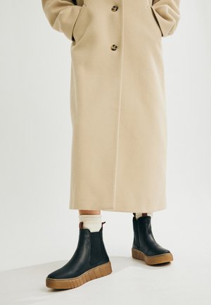 Personne portant un long manteau beige avec les mains dans les poches, des bottines noires avec des semelles épaisses marron, et des chaussettes blanches visibles au-dessus des bottines.