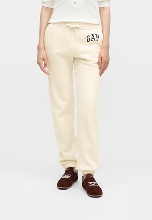 Osoba nosiaca krémové tepláky s logom „GAP“ na ľavom boku a tmavočervené tenisky s bielymi šnúrkami, stojaca na jednoduchom bielom pozadí.