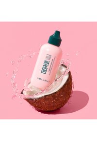 Roze fles van het haarelixer "Coco & Eve Like a Virgin" spat uit een doormidden gesneden kokosnoot op een roze achtergrond.