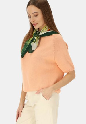 Kobieta ubrana w brzoskwiniowy, dzianinowy top z krótkim rękawem, kremowe spodnie z ręką w kieszeni oraz zielony kwiecisty szalik związany wokół szyi.