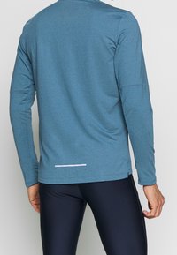 Maglia sportiva a maniche lunghe di colore blu, realizzata in tessuto strutturato, con collo rotondo e una striscia riflettente bianca sull'orlo posteriore.