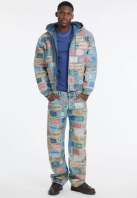 Młody mężczyzna w dżinsowej kurtce i dżinsach ozdobionych kolorowymi naszywkami z logo Guess, w niebieskiej koszuli i brązowych butach, stojący z rękami w kieszeniach.