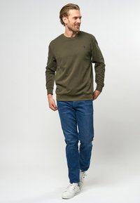 Olijfgroene katoenen sweatshirt met geribbelde manchetten en onderrand, gecombineerd met blauwe jeans en witte sneakers, gedragen door een man die loopt.