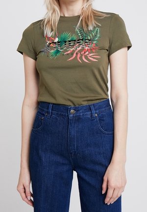 Person wearing olive green t-shirt with colorful tropical leaves and "GUESS Los Angeles" text, paired with high-waisted blue jeans.