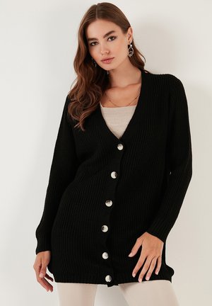 Black knitted cardigan with a deep V-neck and long sleeves; features large white buttons down the front and ribbed texture throughout.