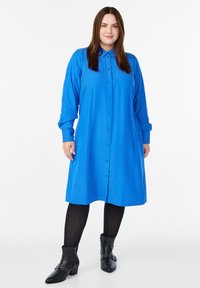 Kobieta ubrana w błękitną sukienkę w paski z długim rękawem, czarne rajstopy i czarne botki z klamrami, stojąca na białym tle.