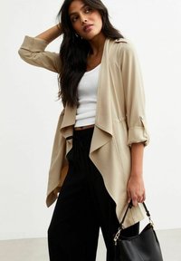 Giacca beige leggera aperta davanti con design drappeggiato, maniche a tre quarti e dettagli con bottoni. Abbinata a pantaloni neri e una borsa.