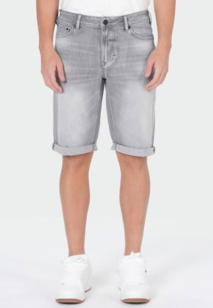 Mann trägt hellgraue Jeans-Shorts mit hochgekrempelten Beinen und weißen Sneakers, steht vor einem einfarbigen hellen Hintergrund.