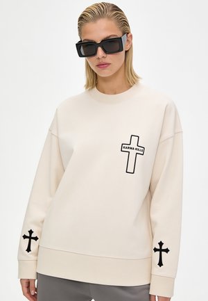 Femme aux cheveux blonds courts portant de grandes lunettes de soleil noires et un sweat-shirt beige avec des motifs croix noirs et le texte "Karma Kills".