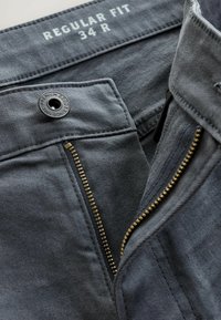 Pelēki džinsi, parastā piegriezuma. Iezīmēts ar misiņa rāvējslēdzēju, melnu pogu ar uzrakstu "Premium Goods" un kontrastējošiem dūrieniem jostasvietā.