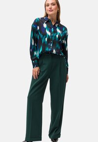 Blusa di satin con stampa astratta blu, verde e nera e maniche lunghe, abbinata a pantaloni verdi a vita alta con pince.