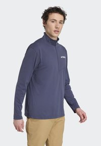 Námornícka modrá, ľahká košeľa s dlhým rukávom, s polo zipsom, vysokým golierom a logom na hrudi. Hladká textúra a pohodlný strih.