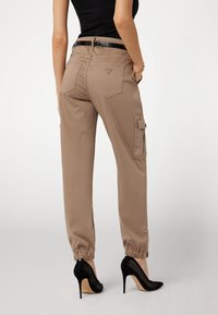 Pantalons cargo kaki avec des poignets élastiques, poche latérale et poches arrière. Portés avec des escarpins noirs à talons aiguilles, mettant en valeur une silhouette élancée.