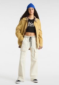 Geel corduroy jack, zwart cropped t-shirt, crèmekleurige cargobroek met zakken, zwarte trekkoord, en blauwe gebreide beanie, zwart-witte sneakers.