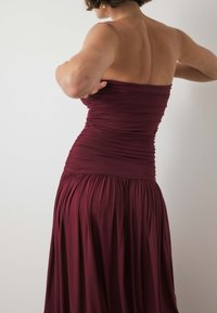 Burgundia õlgetu kleit, millel on väänatud kehaosa ja voolav volditud seelik. Valmistatud pehmest kangast, millel on sile tekstuur.