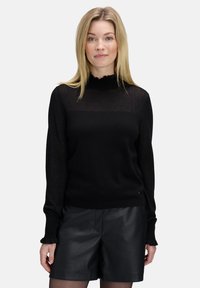 Zwarte coltrui met ruches aan de mouwen, gemaakt van een zachte, gestructureerde stof. Gecombineerd met zwarte leren shorts voor contrast.