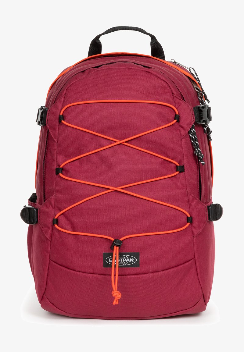 Punainen reppu, jossa on oranssi bungee-köysisuunnittelu, valmistettu kestävästä kankaasta. Siinä on sivutaskut ja kantokahva helppoa kuljettamista varten.
