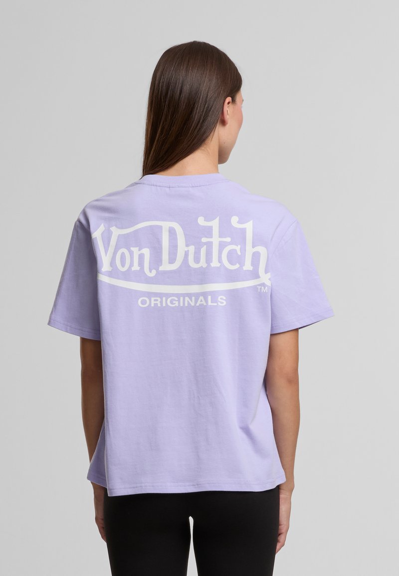 Mladá žena s dlouhými hnědými vlasy, oblečená v světle fialovém tričku Von Dutch Originals, viděná zezadu na jednolitém pozadí.