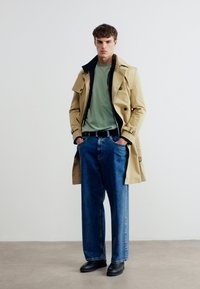 Νεαρός άνδρας με μπεζ καμπαρντίνα πάνω από ανοιχτό πράσινο πουκάμισο και άνετο μπλε τζιν παντελόνι, στέκεται μπροστά σε λευκό φόντο.