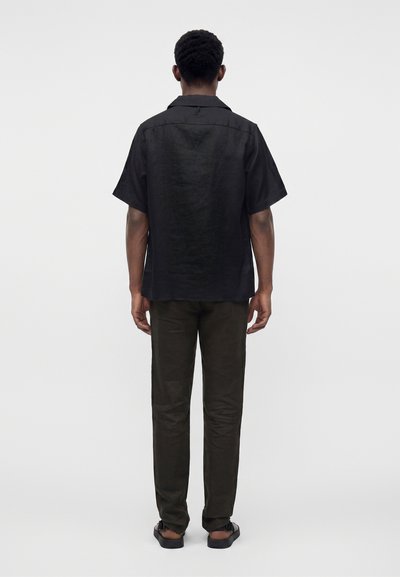 Homme debout, de dos, portant une chemise noire à manches courtes avec boutons, un pantalon foncé et des sandales noires sur un fond uni.