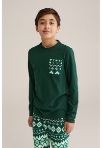 Tricou cu mânecă lungă de culoare verde închis, cu un buzunar cu model, combinat cu pantaloni verzi și albi cu modele, care prezintă forme de diamant și designuri geometrice.