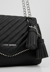 Borsa a tracolla nera in ecopelle trapuntata con catena, con dettagli a nappina e hardware in tono argento. Presenta cuciture decorative e borchie in metallo.
