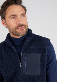 Marineblaues Weste mit strukturiertem Fleece-Vorderteil und glattem Stofftasche. Verfügt über einen hohen Kragen und einen durchgehenden Reißverschluss.