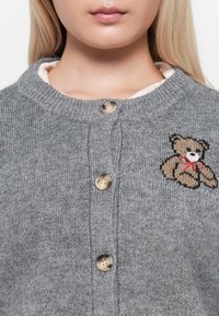 Cardigan grigio lavorato a maglia con scollo rotondo, tre bottoni e un orsetto ricamato con un nastro rosso sul lato sinistro.