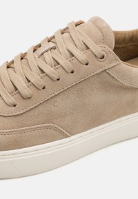 Beige suede sneaker with round toe, flat white rubber sole, and contrasting brown laces. Features embossed logo on side for detail.