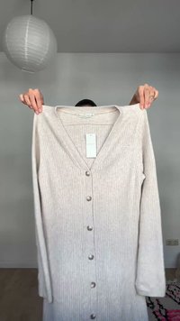 Cardigan beige côtelé à boutons avec manches longues, porté par une personne devant un mur gris avec une lampe ronde blanche suspendue au-dessus.