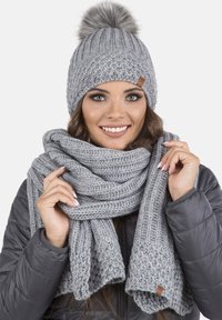 Berretto di maglia grigio con pom-pom e sciarpa abbinata, entrambi con un motivo testurizzato. La sciarpa è avvolta elegantemente, con dettagli in pelle visibili.