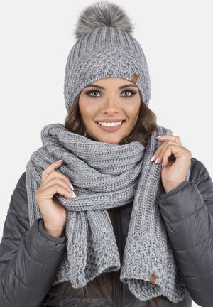 Berretto di maglia grigio con pom-pom e sciarpa abbinata, entrambi con un motivo testurizzato. La sciarpa è avvolta elegantemente, con dettagli in pelle visibili.
