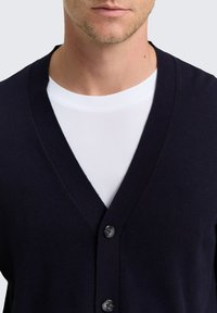 Marinefarbener V-Ausschnitt-Cardigan aus feinem Strickmaterial, mit fünf Knopfverschlüssen, einem strukturierten Stoff und getragen über einem weißen T-Shirt.
