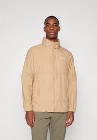 Veste beige légère avec un col montant, fermeture éclair à l'avant et poignets élastiques. Présente un logo sur la poitrine et une finition légèrement texturée.