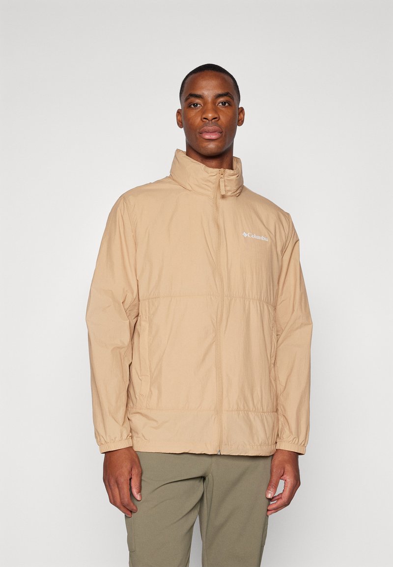 Veste beige légère avec un col montant, fermeture éclair à l'avant et poignets élastiques. Présente un logo sur la poitrine et une finition légèrement texturée.