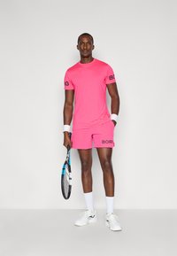 Lyse rosa treningsskjorte og shorts med svart "BORG"-logo. Spiller holder en tennisracket, iført hvite sokker og sneakers.