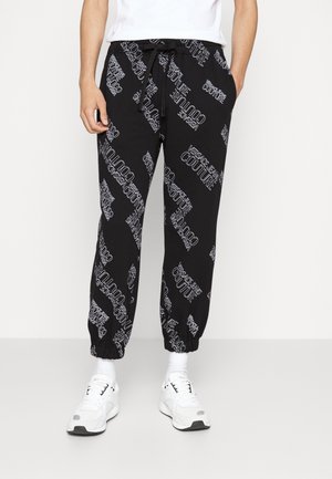 Czarne spodnie dresowe z białym nadrukiem "Versace Jeans Couture", elastyczne ściągacze, noszone z białymi trampkami i białą koszulką.