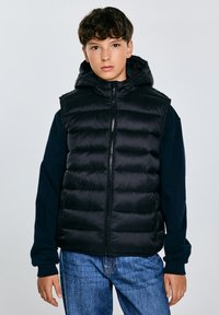Schwarze Pufferweste mit Kapuze, mit gestepptem Design, Reißverschluss und seitlichen Taschen, kombiniert mit einem navy blauem Langarmhemd und blauen Jeans.