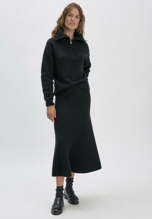 Schwarzer Strickpullover mit einem Viertel-Reißverschluss-Kragen kombiniert mit einem passenden gerippten Midikleid. Glatte Textur, taillierter Schnitt, ausgestellter Saum. Schwarze Schuhe.