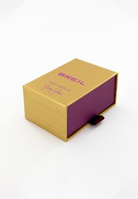 Gul låda med en djup lila insida, med "BREIL" i fet rosa text och ett snöre för att öppna den. Kompakt och rektangulär design.