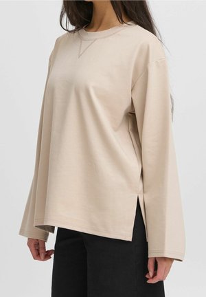 Beżowa bluzka z długim rękawem i okrągłym dekoltem z bocznymi rozcięciami, noszona z czarnymi spodniami, pokazana na osobie od ramion do bioder.