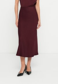 Jupe midi bordeaux, fabriquée en tissu lisse. Taille cintrée avec un ourlet évasé, associée à des talons noirs pointus pour le contraste.