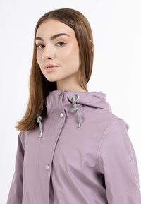 Chaqueta impermeable morada con un alto cuello, cordones grises, botones a presión y detalles reflectantes. Textura suave y diseño ajustado.