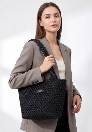 Jeune femme en blazer gris tenant un grand sac cabas noir tissé avec une bandoulière sur fond blanc.